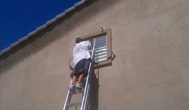 A window cleaner on a ladder working on a small second-story window under a tile roof