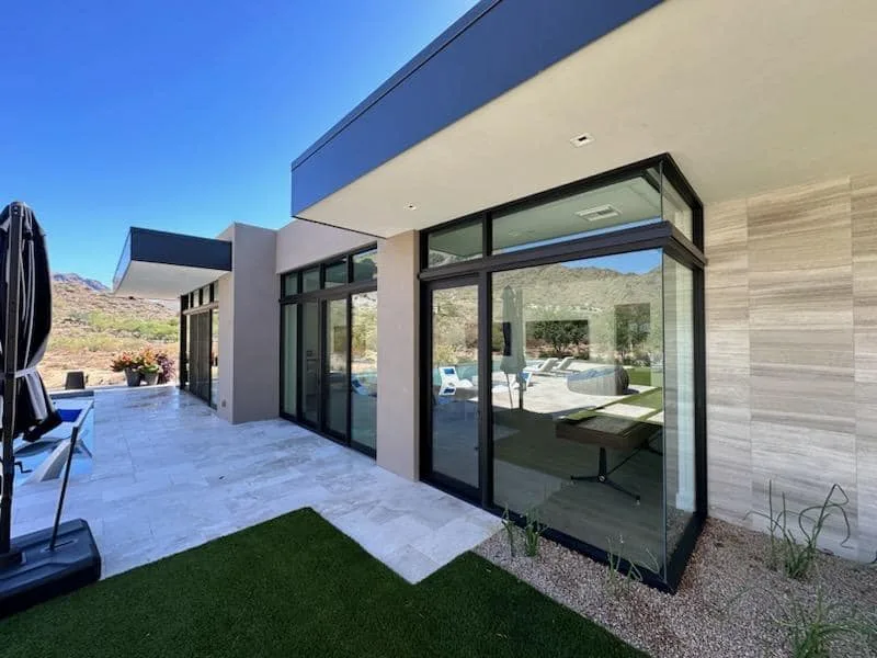 A modern desert home with floor-to-ceiling glass walls opening onto a patio, with mountains behind